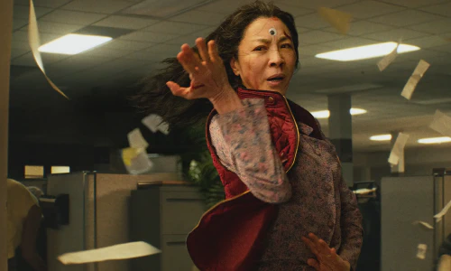 A screencap from the movie Everything Everywhere All at Once showing Evelyn (played by Michelle Yeoh) standing in a fighting pose in the middle of a sea of office cubicles. A single googly eye is stuck to her forehead.