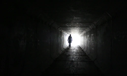 Black and white photo of a person's silhouette standing in a square hallway. The opposite end of the hallway is drenched in a blinding white light, while the nearer end is shrouded in shadow.