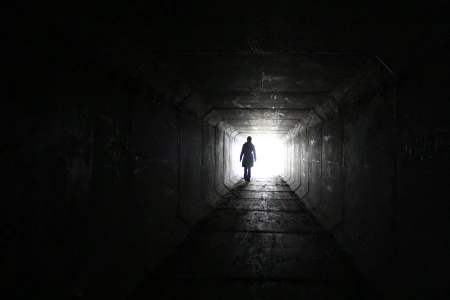 Black and white photo of a person's silhouette standing in a square hallway. The opposite end of the hallway is drenched in a blinding white light, while the nearer end is shrouded in shadow.